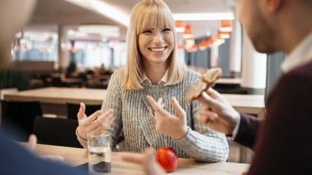 Kollegin in der Kaufland-Kantine im Gespräch mit ihren Kollegen. Vor ihr auf dem Tisch: ein Wasserglas und ein Apfel.