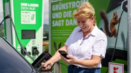 Frau lädt E-Auto mit Ökostrom von Kaufland und schaut lachend auf ihr Handy.