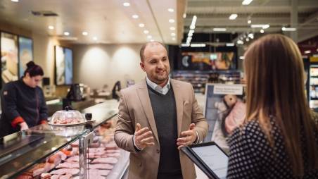 Kaufland-Filialleiter im Gespräch mit einer Mitarbeiterin, die ein Tablet in den Händen hält. Im Hintergrund eine Wursttheke.