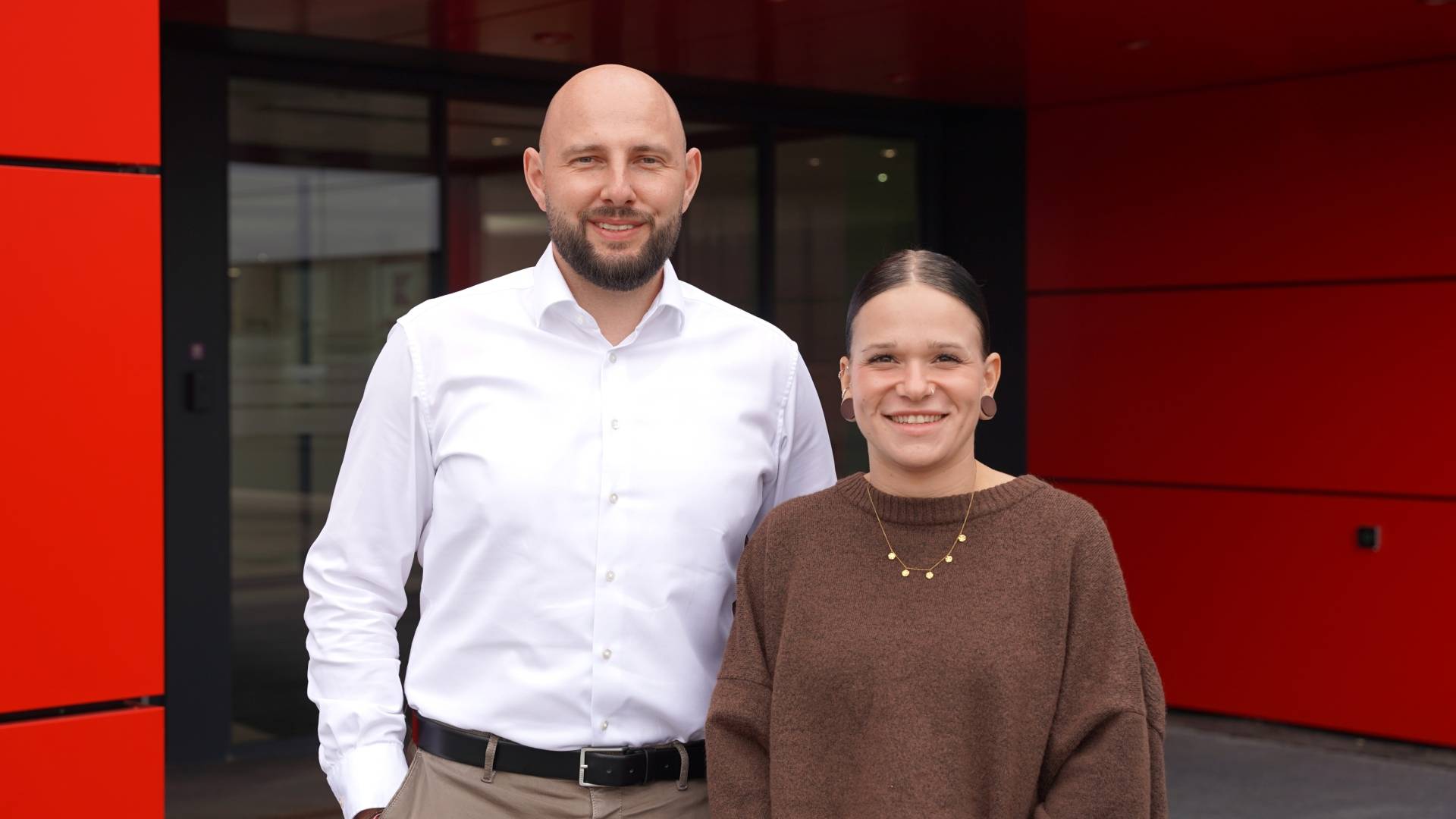 Zwei Mitarbeiter aus dem Zentraleinkauf bei Kaufland International stehen vor einem Bürogebäude und lächeln in die Kamera.